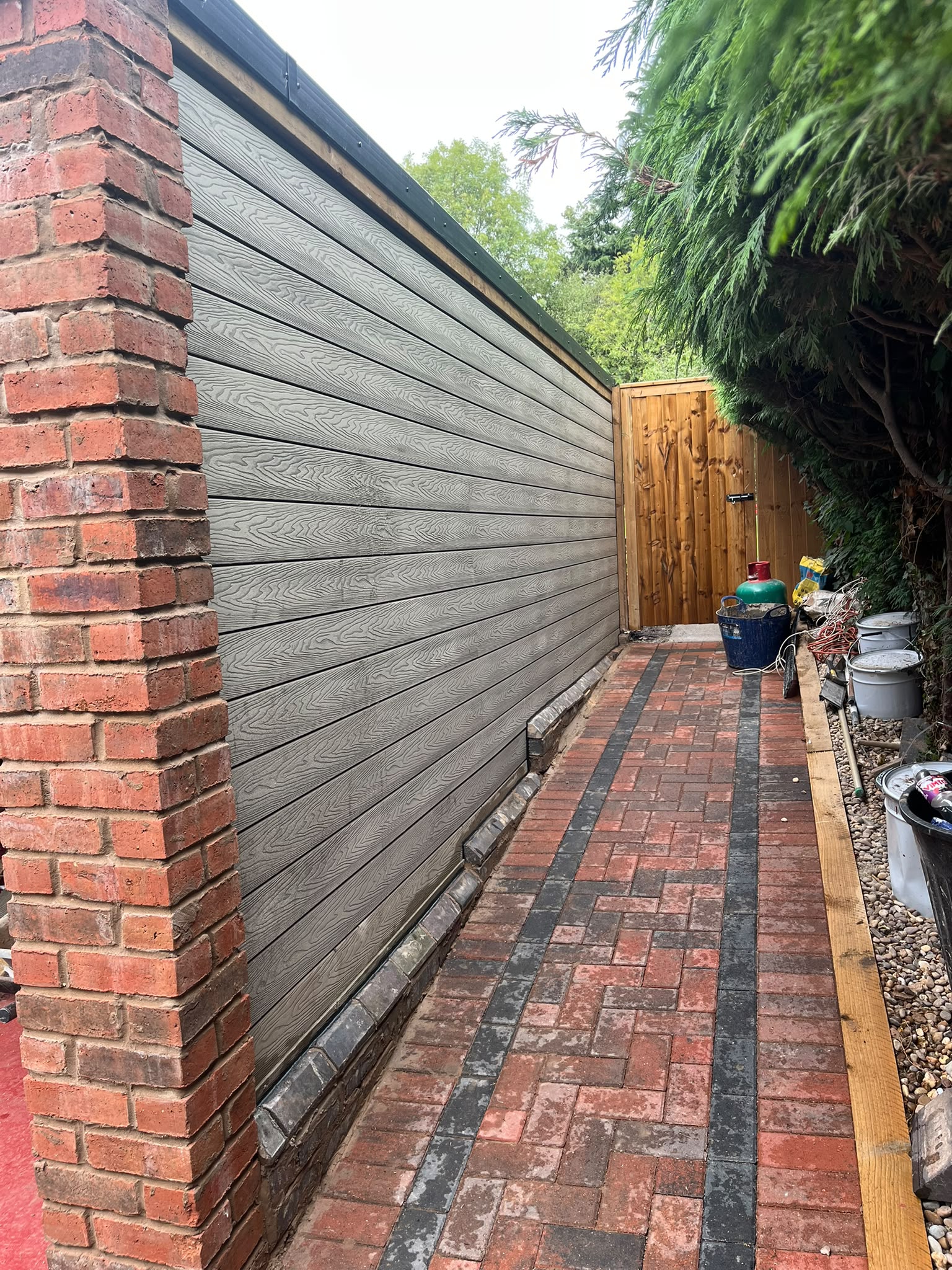 Red and black brick paving in a narrow alleyway beside grey cladding and a timber gate